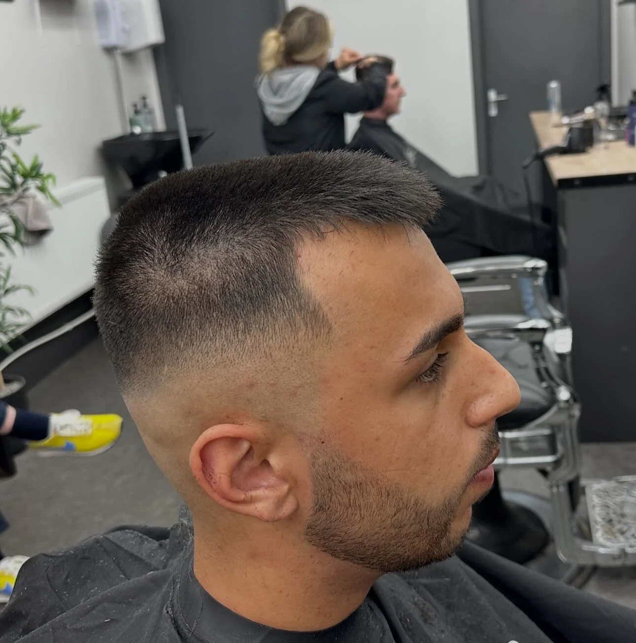 Short crew cut with a crisp fade, barber at work in the background